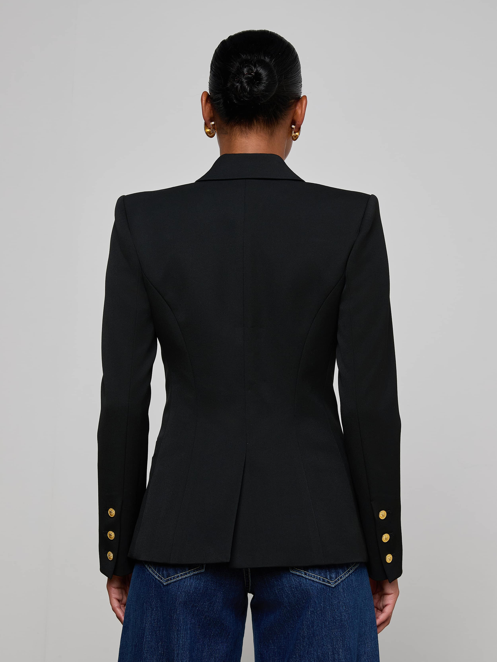 A woman stands facing away, wearing the Kyomi Double-Breasted Blazer with gold buttons on the sleeves and blue jeans. Her hair is styled in a neat bun against a plain, light-colored background.