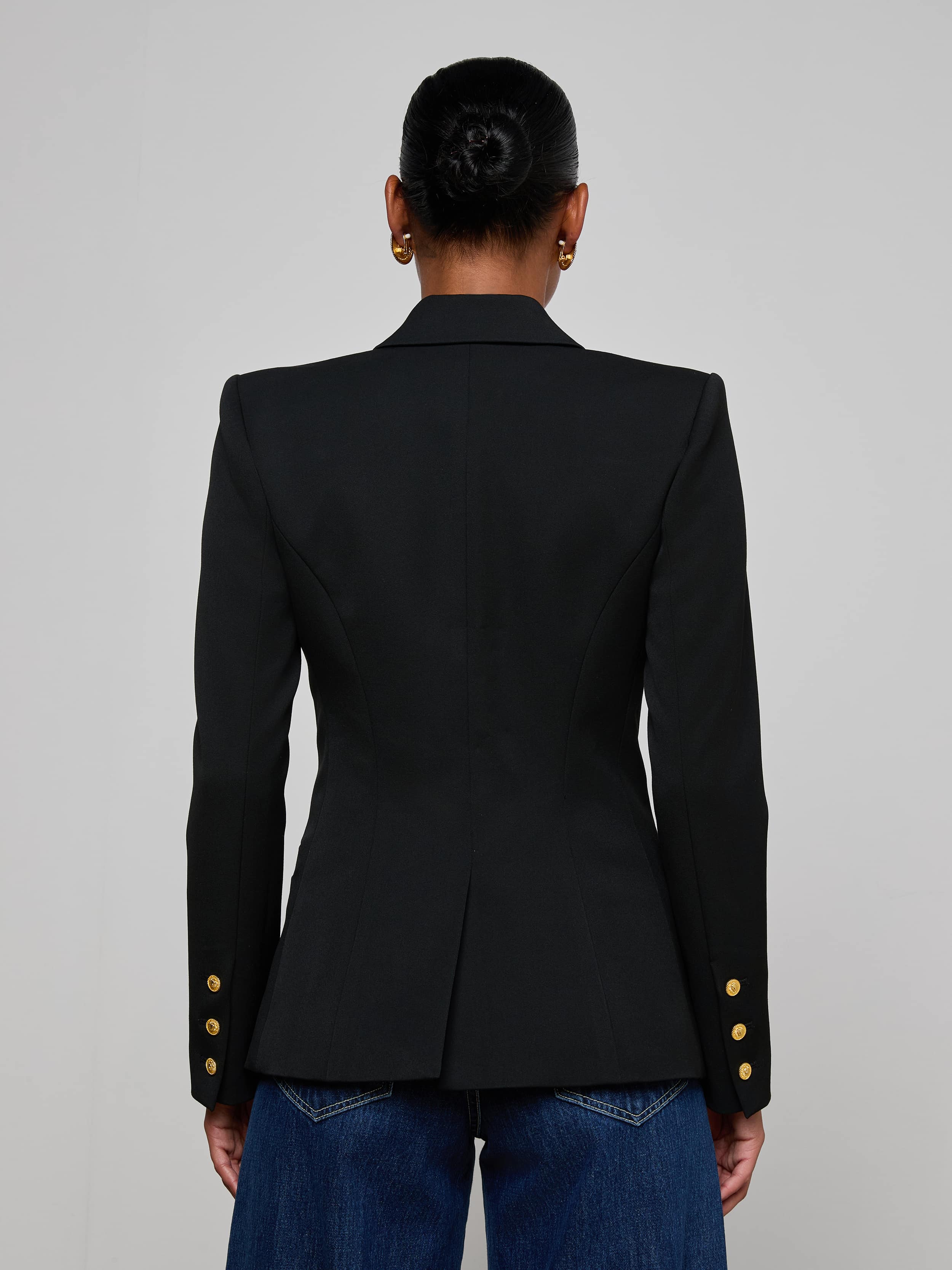 A woman stands facing away, wearing the Kyomi Double-Breasted Blazer with gold buttons on the sleeves and blue jeans. Her hair is styled in a neat bun against a plain, light-colored background.