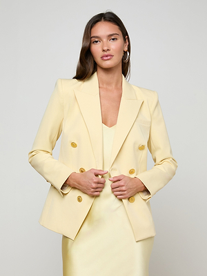 A woman with long brown hair wears the Kyomi Double-Breasted Blazer in light yellow over a matching dress, standing against a plain light background and looking at the camera with a neutral expression.