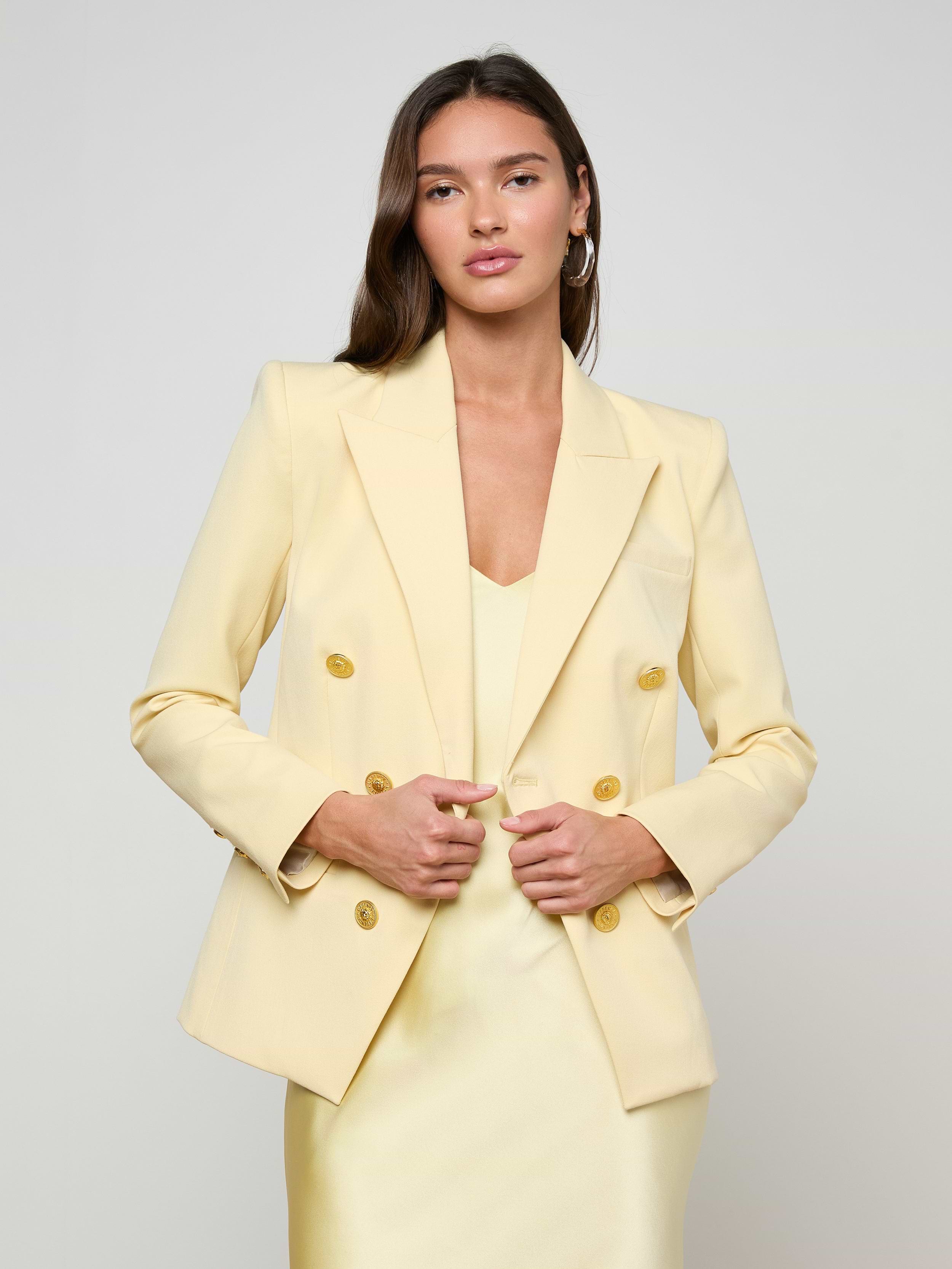 A woman stands facing forward against a plain white background, hands holding the front of her Kyomi Double-Breasted Blazer in yellow, styled over a matching dress.