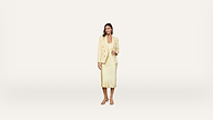 A woman stands against a plain white background wearing a light yellow blazer and matching midi dress with strappy sandals.