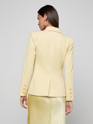 A woman stands with her back to the camera, wearing the Kyomi Double-Breasted Blazer in pale yellow with gold sleeve buttons and a matching shiny skirt, set against a plain light gray background.