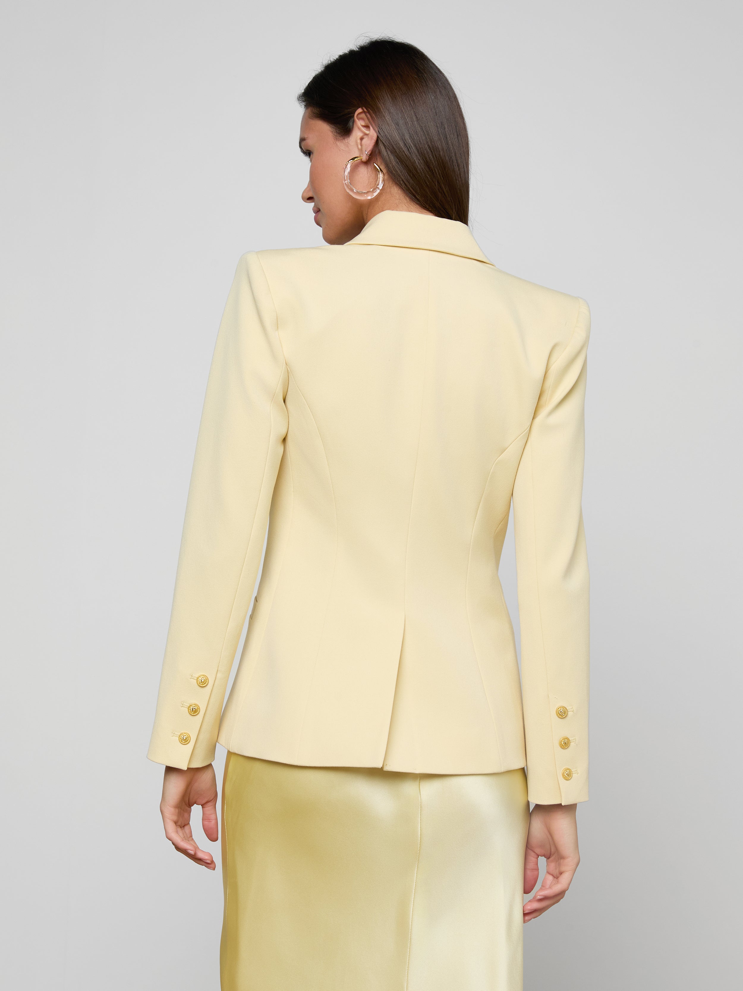 A woman is shown from the back wearing the Kyomi Double-Breasted Blazer in yellow with gold buttons and a matching satin skirt, standing against a plain off-white background.