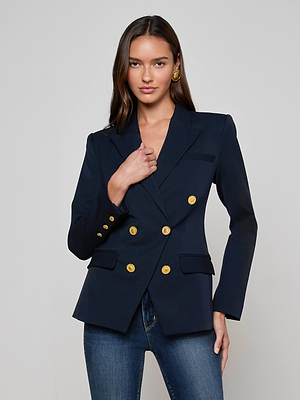 A woman with long brown hair wears the L'AGENCE Kyomi Double-Breasted Blazer with gold buttons and blue jeans, standing against a plain light gray background. She touches her lapel and looks at the camera.