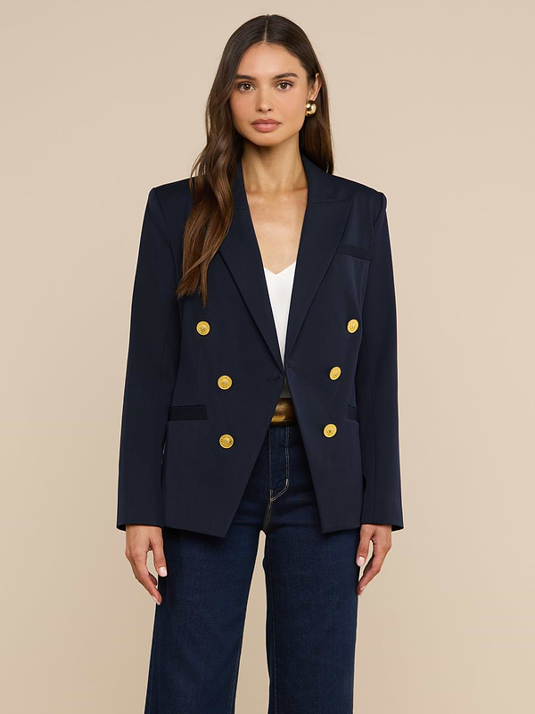 Su uno sfondo beige, una donna dai lunghi capelli castani e dall’espressione neutra indossa il blazer doppiopetto Kyomi in blu navy con bottoni dorati, abbinato a un top bianco e a jeans blu scuro.
