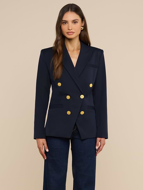 Una donna dai lunghi capelli castani indossa il blazer doppiopetto Kyomi, caratterizzato da bottoni dorati, sopra un top scuro e dei jeans, su uno sfondo beige, guardando dritto verso la fotocamera.