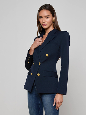 A woman with long brown hair, wearing the L'AGENCE Kyomi Double-Breasted Blazer with gold buttons, blue jeans, and gold earrings, stands against a light gray background facing forward and slightly smiling.