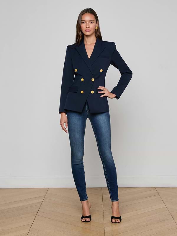 A woman stands against a plain white background, wearing sunglasses, the Kyomi Double-Breasted Blazer in midnight navy with gold buttons, a white top, dark wide-leg jeans, and tan pointed-toe shoes.