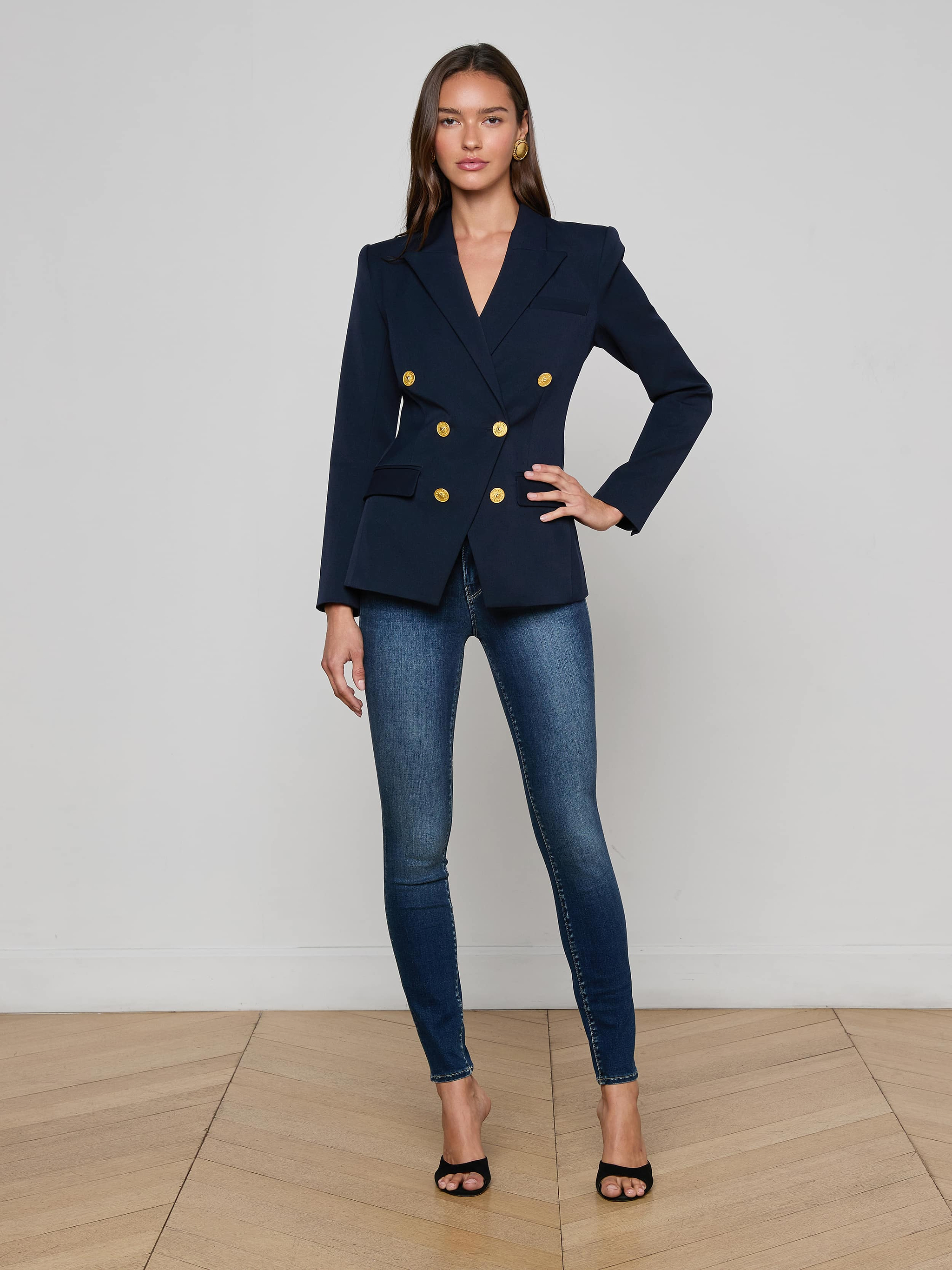 A woman stands against a plain white background, wearing sunglasses, the Kyomi Double-Breasted Blazer in midnight navy with gold buttons, a white top, dark wide-leg jeans, and tan pointed-toe shoes.