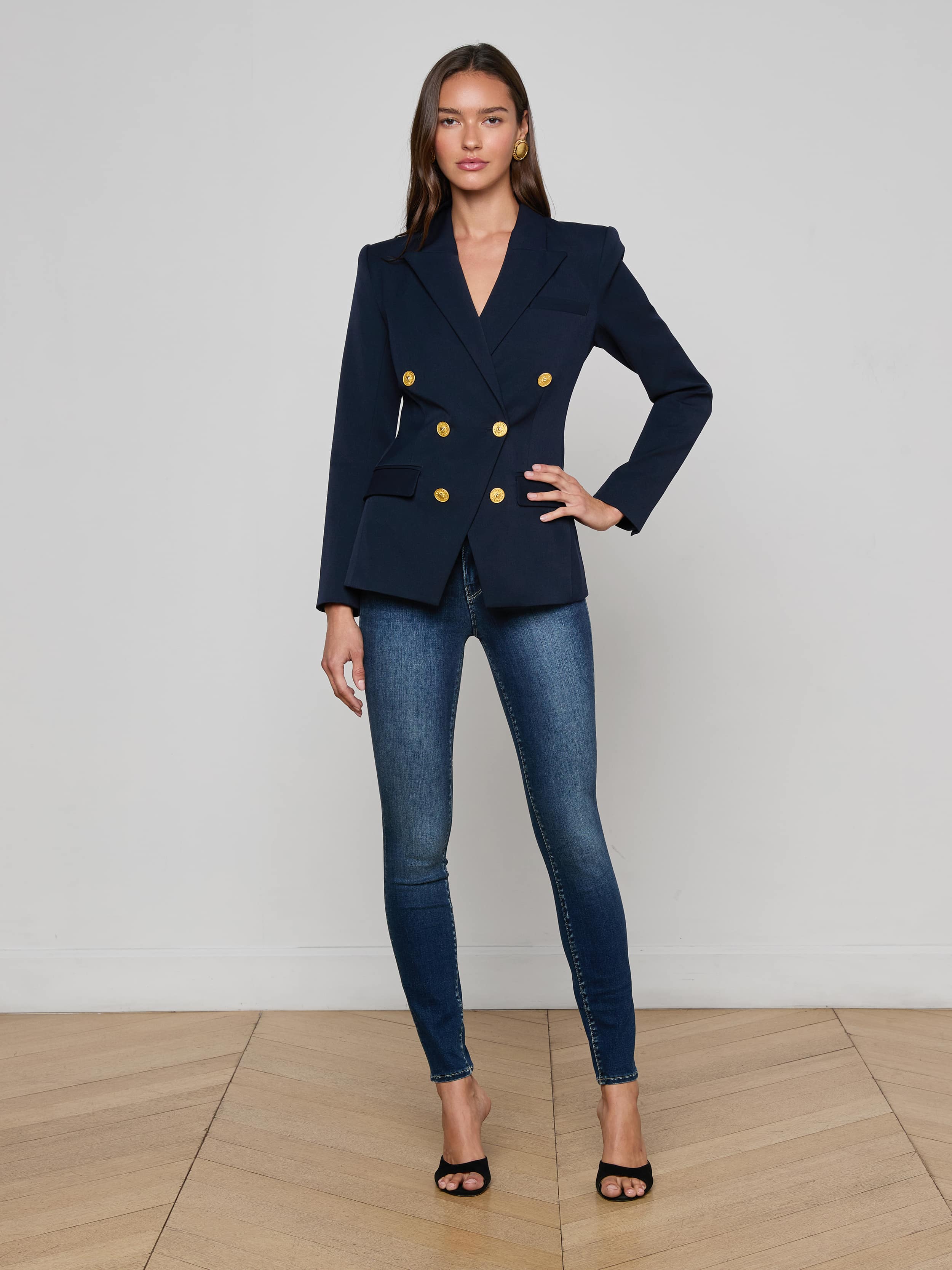 A woman stands against a plain white background, wearing sunglasses, the Kyomi Double-Breasted Blazer in midnight navy with gold buttons, a white top, dark wide-leg jeans, and tan pointed-toe shoes.