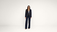 A person wearing sunglasses, a navy blazer with gold buttons, a white shirt, and wide-leg navy pants stands against a plain white background.