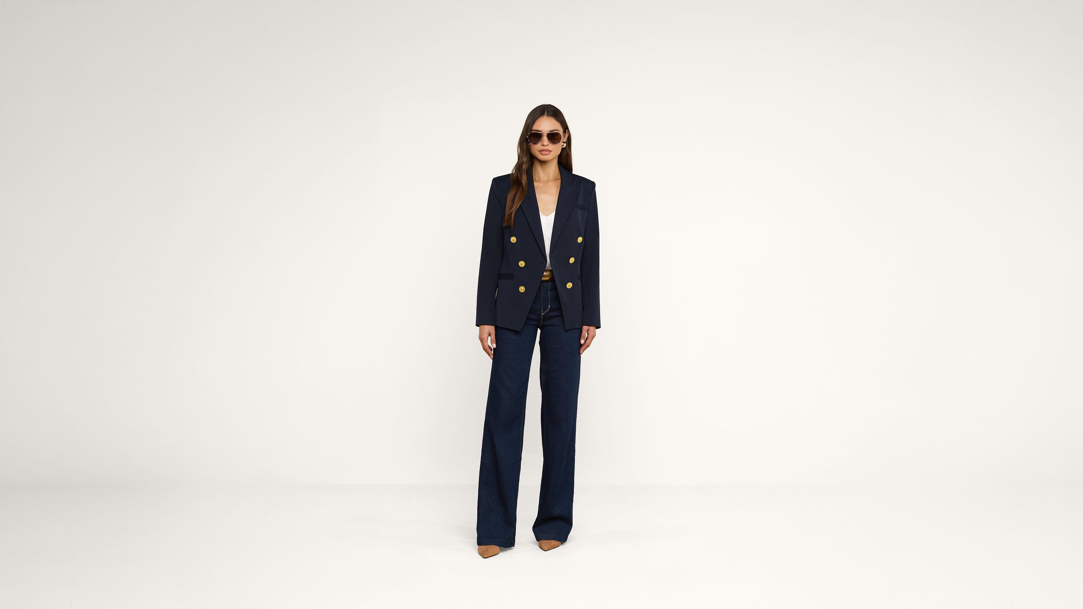 A person wearing sunglasses, a navy blazer with gold buttons, a white shirt, and wide-leg navy pants stands against a plain white background.