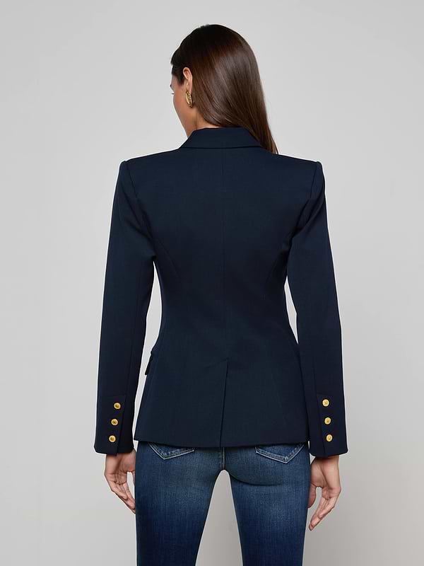 A woman with long brown hair is seen from the back, wearing the Kyomi Double-Breasted Blazer in midnight navy with gold buttons on the sleeves and paired with dark blue jeans, standing against a plain, light background.