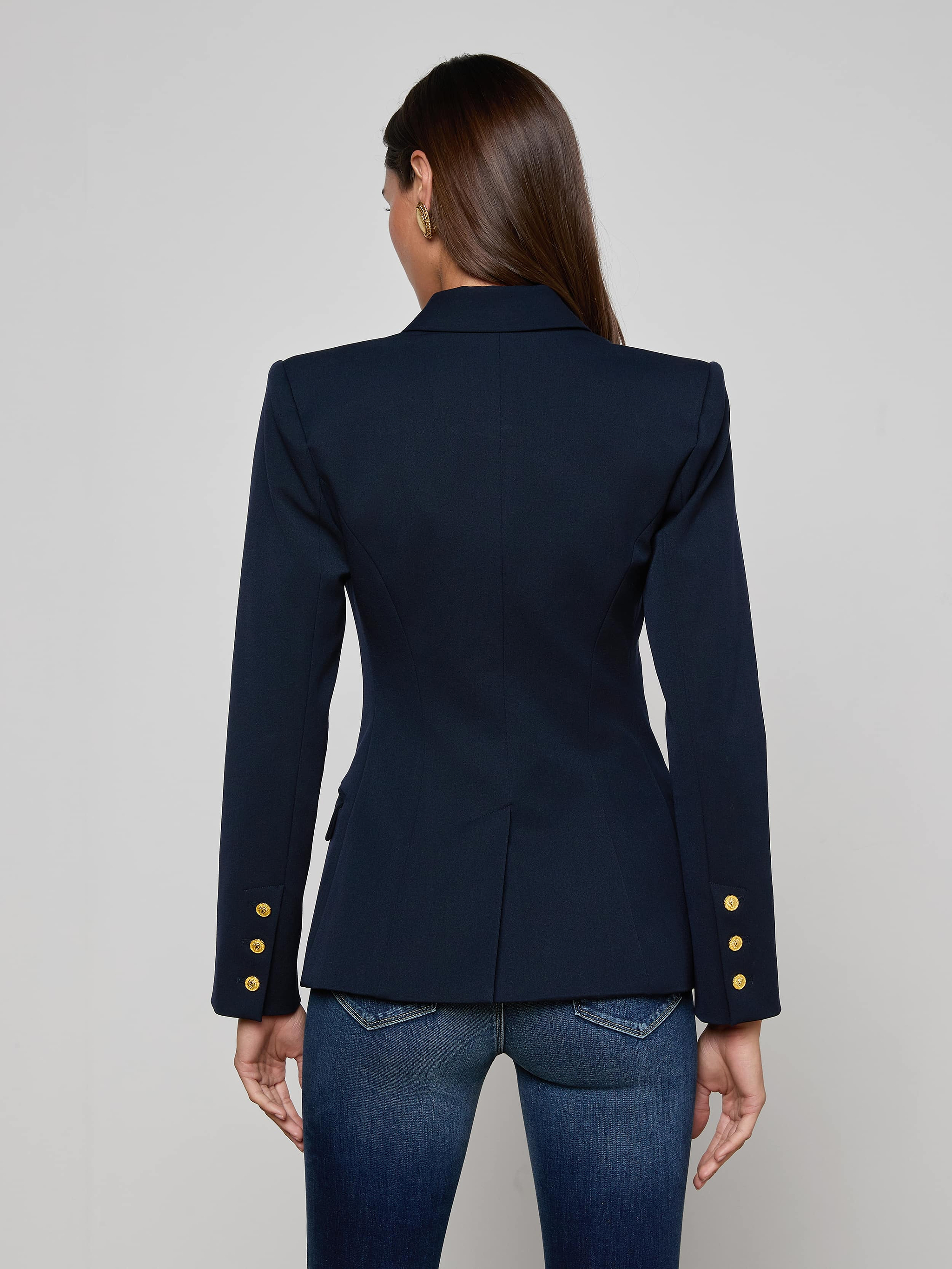 A woman with long brown hair is seen from the back, wearing the Kyomi Double-Breasted Blazer in midnight navy with gold buttons on the sleeves and paired with dark blue jeans, standing against a plain, light background.