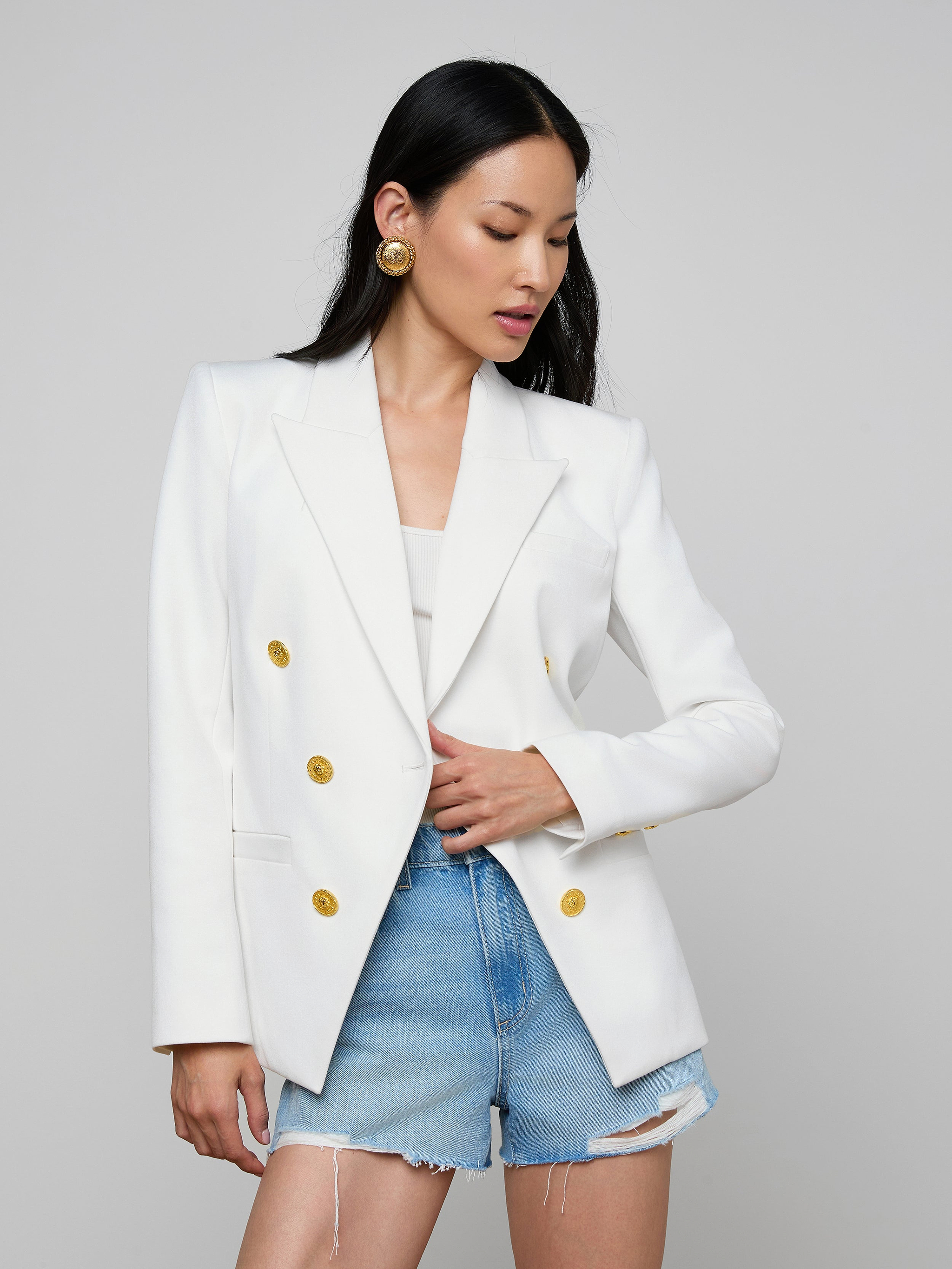 Woman wearing the Kyomi Double-Breasted Blazer in white stretch suit fabric with gold buttons over a white top, paired with high-waisted, frayed denim shorts and gold statement earrings, standing against a light background.