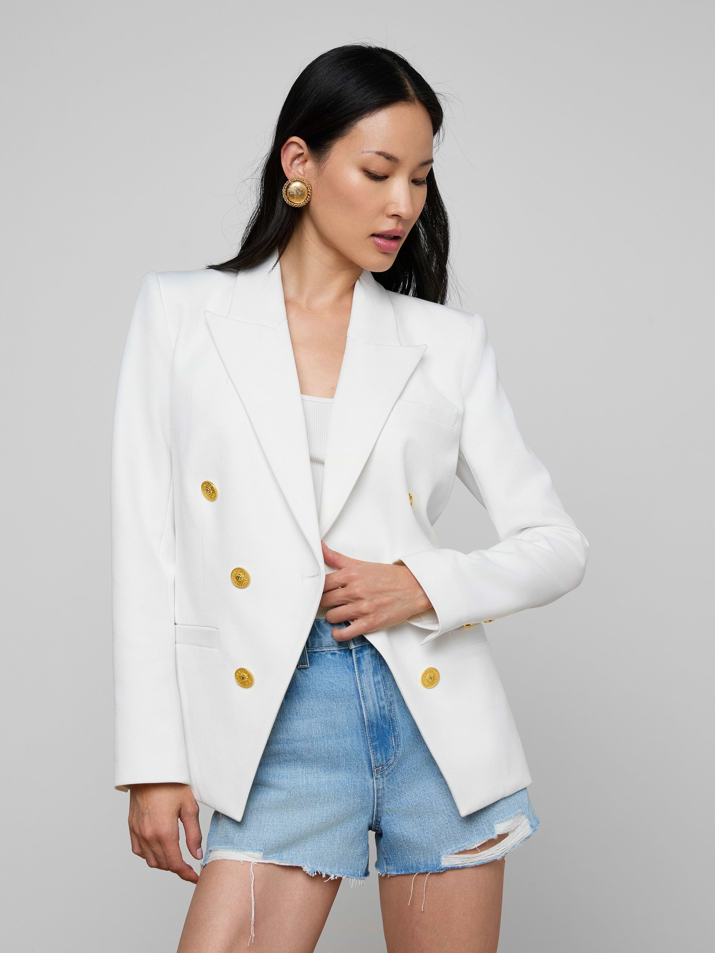 Woman wearing the Kyomi Double-Breasted Blazer in white stretch suit fabric with gold buttons over a white top, paired with high-waisted, frayed denim shorts and gold statement earrings, standing against a light background.