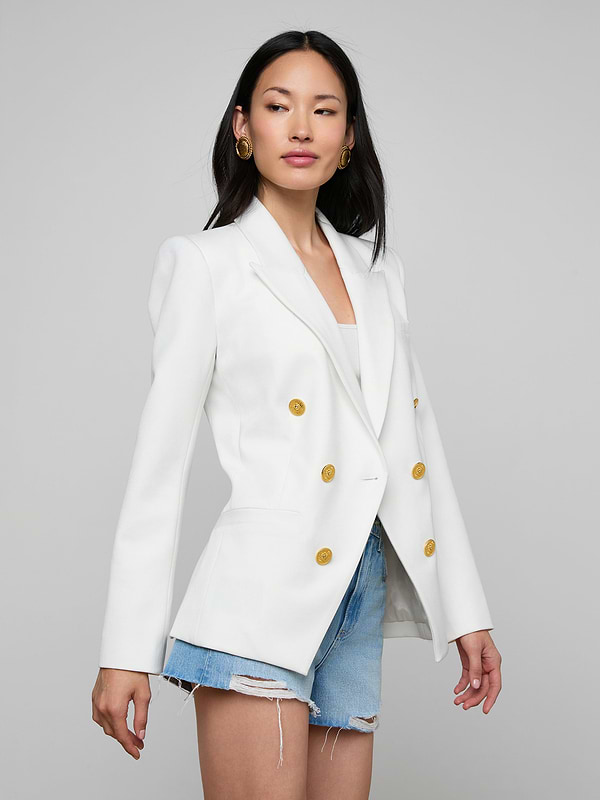 A woman wearing the Kyomi Double-Breasted Blazer in white, crafted from stretch suit fabric, paired with distressed denim shorts, stands against a plain light background.