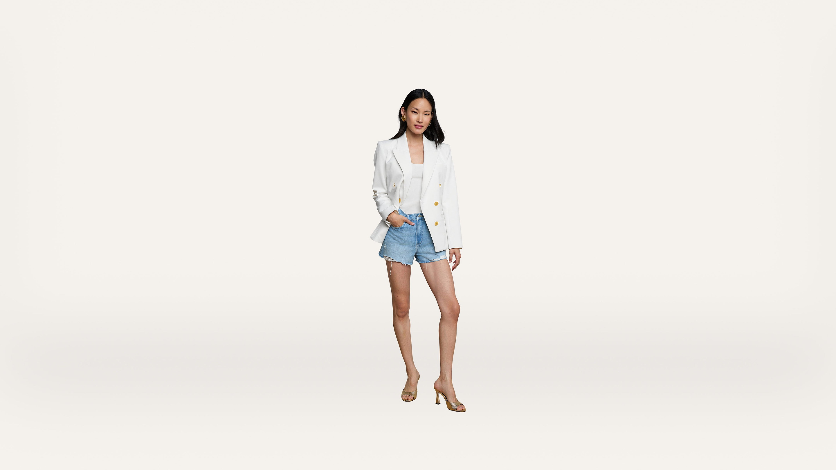 A woman stands confidently wearing a white blazer, white top, denim shorts, and beige heels against a plain white background.