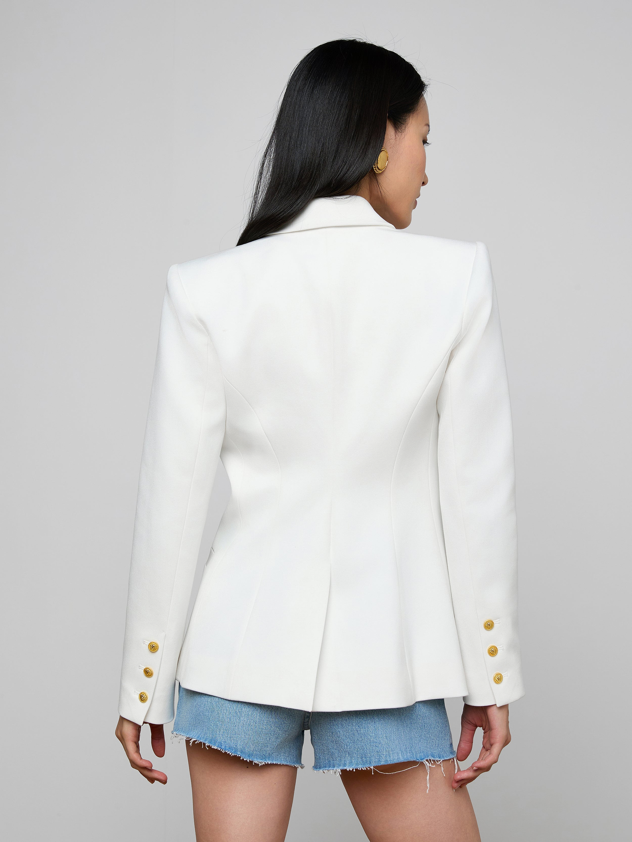 A woman with long black hair is seen from the back, wearing the Kyomi Double-Breasted Blazer—crafted from stretch suit fabric in white with gold buttons—paired with frayed denim shorts against a plain background.