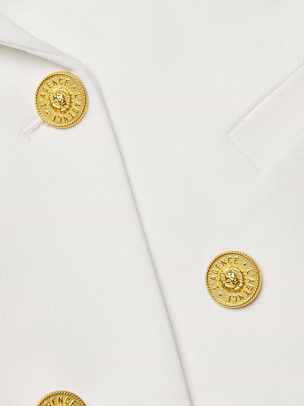 Close-up of the Kyomi Double-Breasted Blazer in white stretch suit fabric, featuring two gold lion’s head-embossed decorative buttons encircled by a textured pattern. The fabric edges are sharply angled.