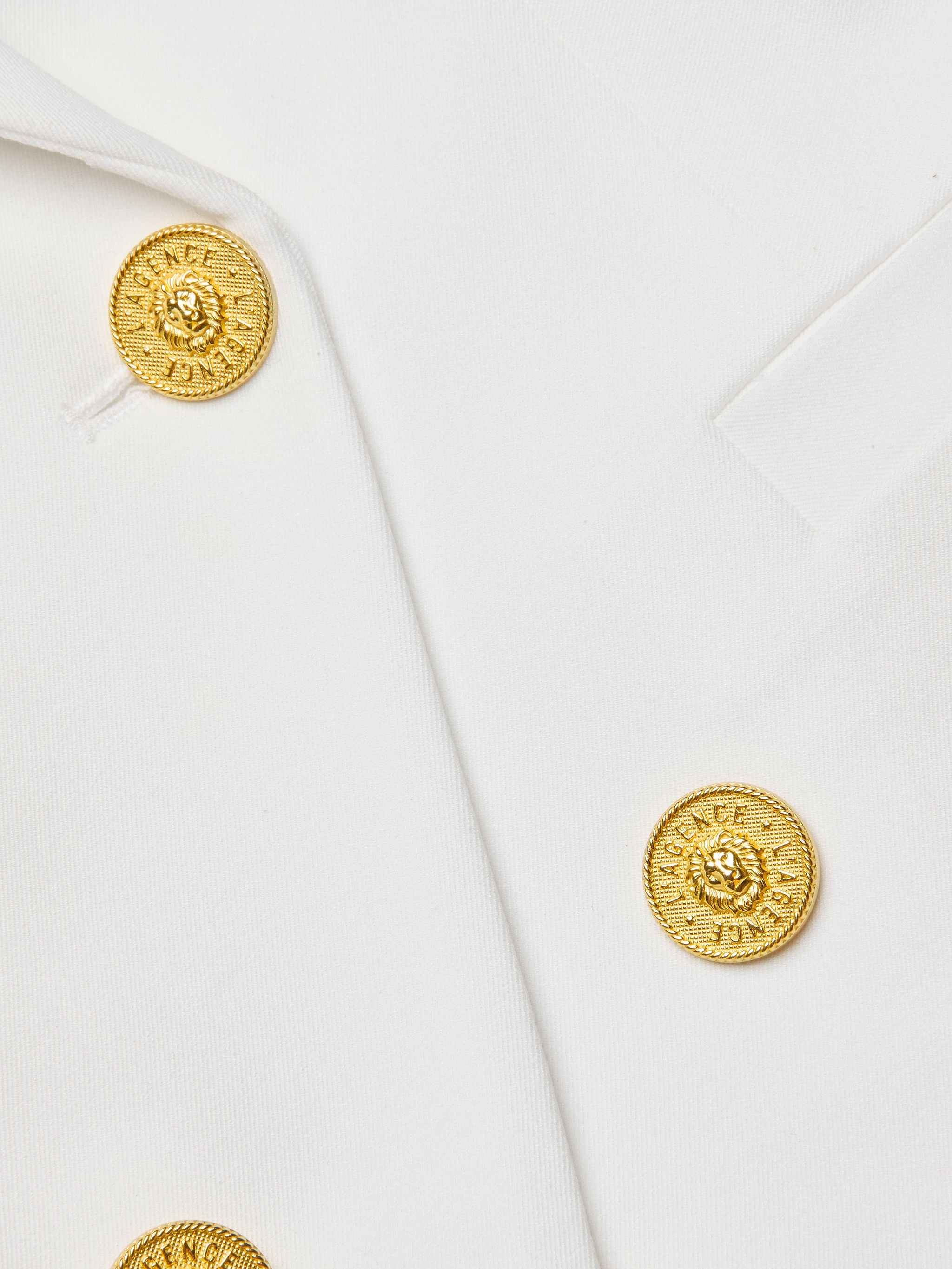 Close-up of the Kyomi Double-Breasted Blazer in white stretch suit fabric, featuring two gold lion’s head-embossed decorative buttons encircled by a textured pattern. The fabric edges are sharply angled.