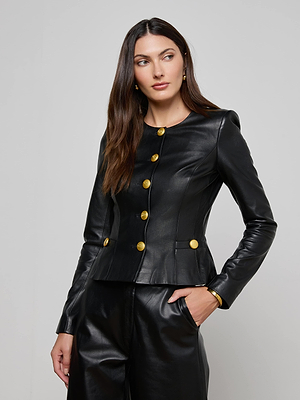 Wearing the Mariella Leather Jacket with gold buttons and matching black leather pants, a woman stands with one hand in her pocket, gazing to the side against a plain gray background.