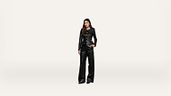 A woman stands against a plain white background wearing a black suit with gold buttons and wide-leg pants, looking directly at the camera.