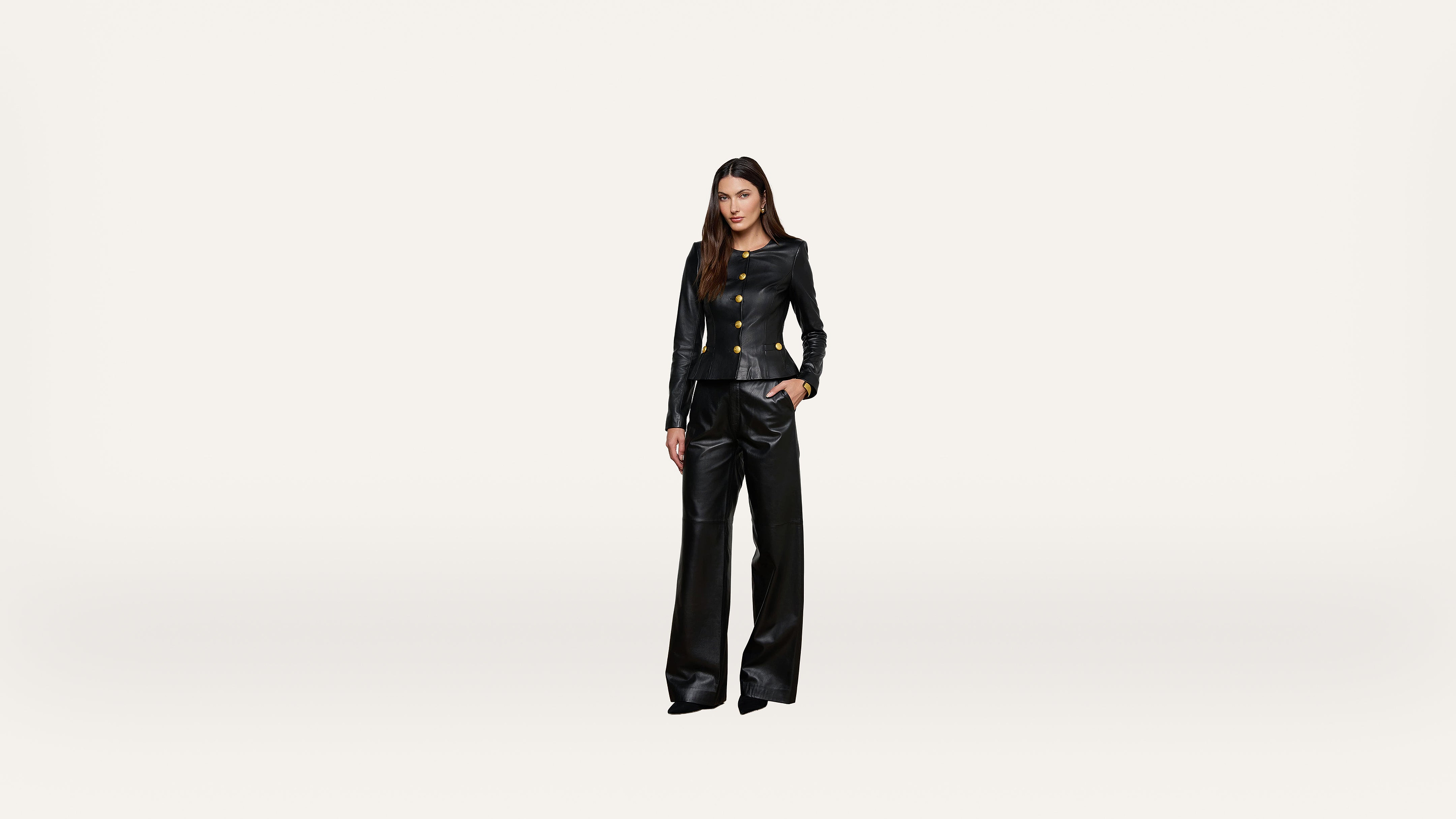 A woman stands against a plain white background wearing a black suit with gold buttons and wide-leg pants, looking directly at the camera.