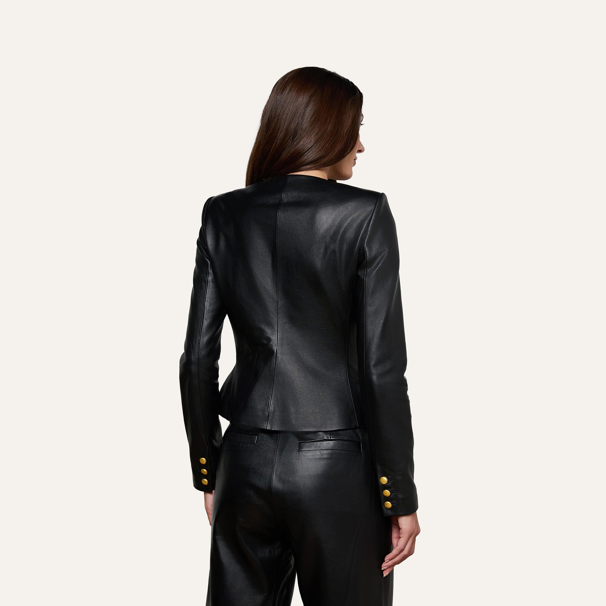 A woman with long brown hair, seen from the back, wears the Mariella Leather Jacket—a fitted collarless style with gold buttons—paired with matching black leather pants, standing against a plain light background.