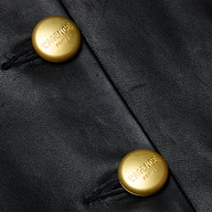 Close-up of two gold buttons on black lambskin leather, each engraved with VAGENU PARIS. The buttons are sewn onto the leather with black thread, showcasing the refined craftsmanship of the Mariella Leather Jacket.