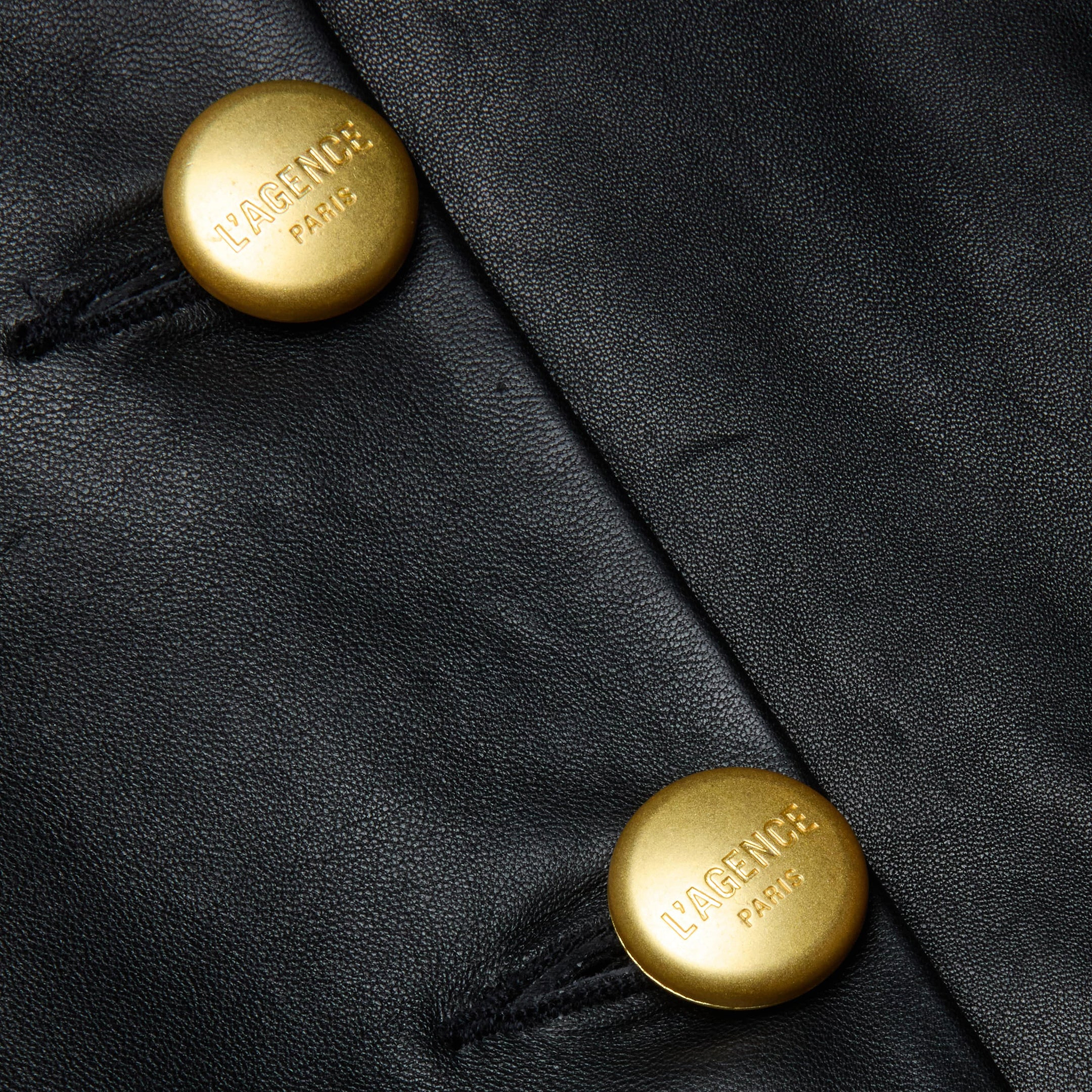 Close-up of two gold buttons on black lambskin leather, each engraved with VAGENU PARIS. The buttons are sewn onto the leather with black thread, showcasing the refined craftsmanship of the Mariella Leather Jacket.