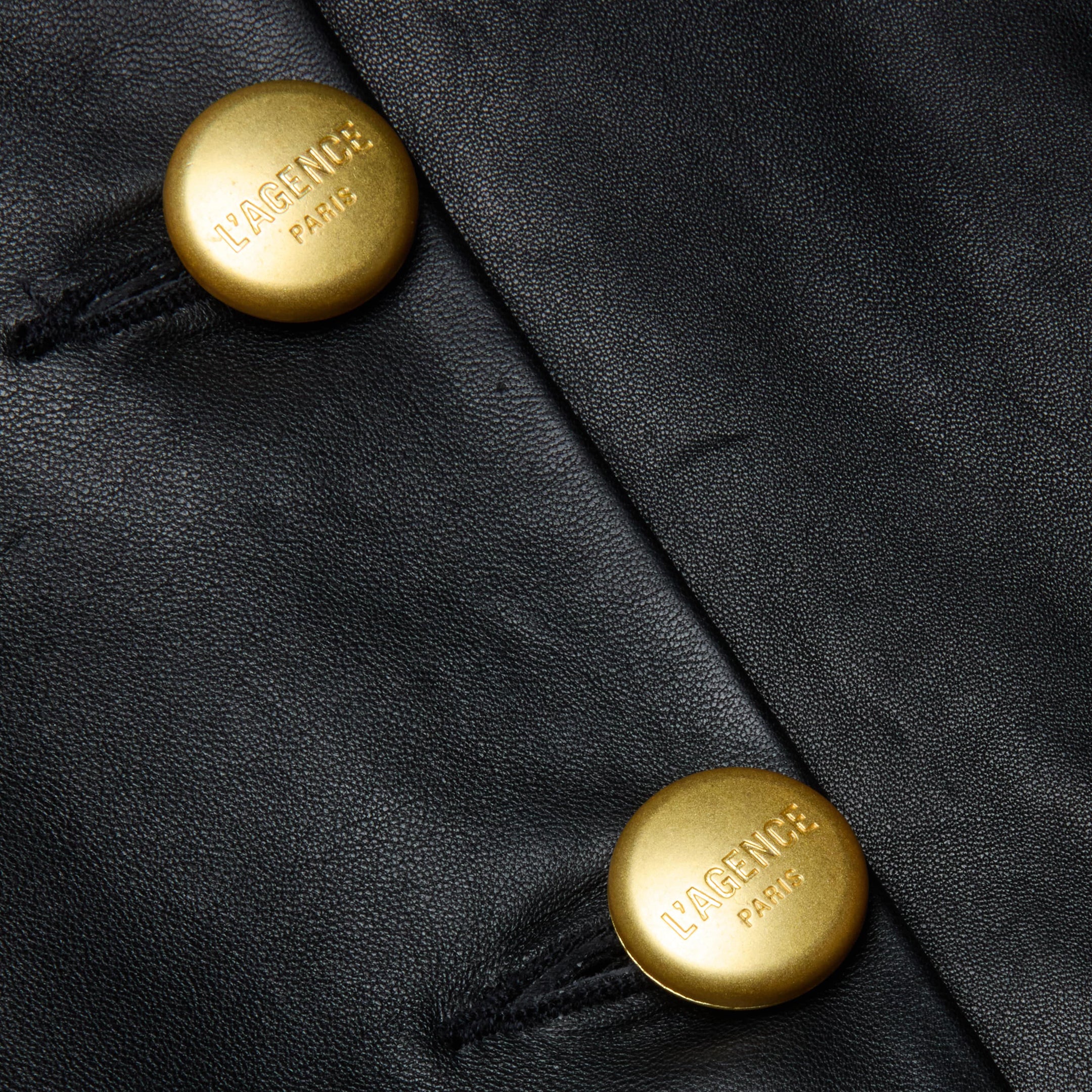 Close-up of two gold buttons on black lambskin leather, each engraved with VAGENU PARIS. The buttons are sewn onto the leather with black thread, showcasing the refined craftsmanship of the Mariella Leather Jacket.