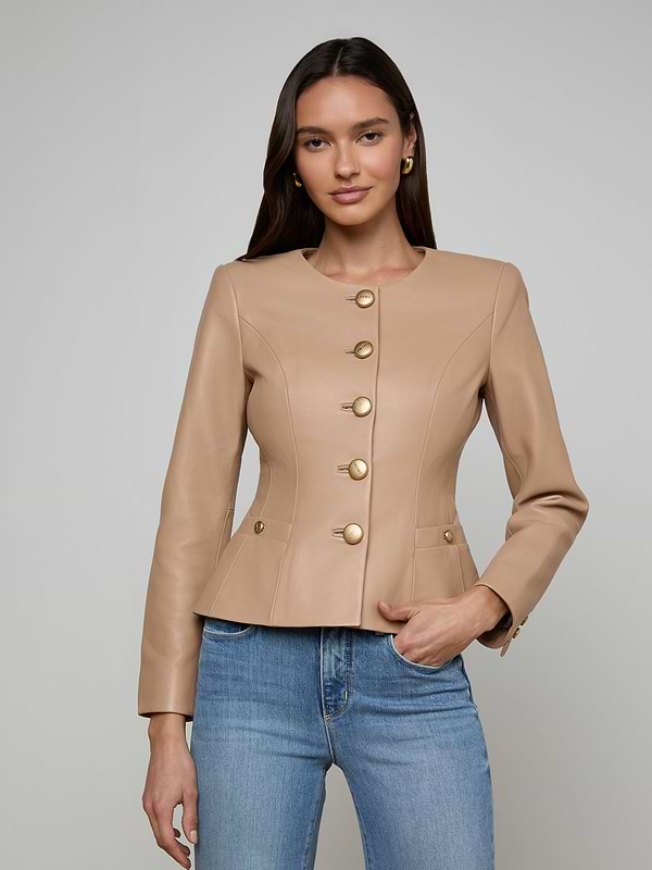 A woman wearing the Mariella Leather Jacket—a fitted, camel-colored chic leather piece with gold buttons—paired with blue jeans, stands against a plain, light background with a neutral expression.