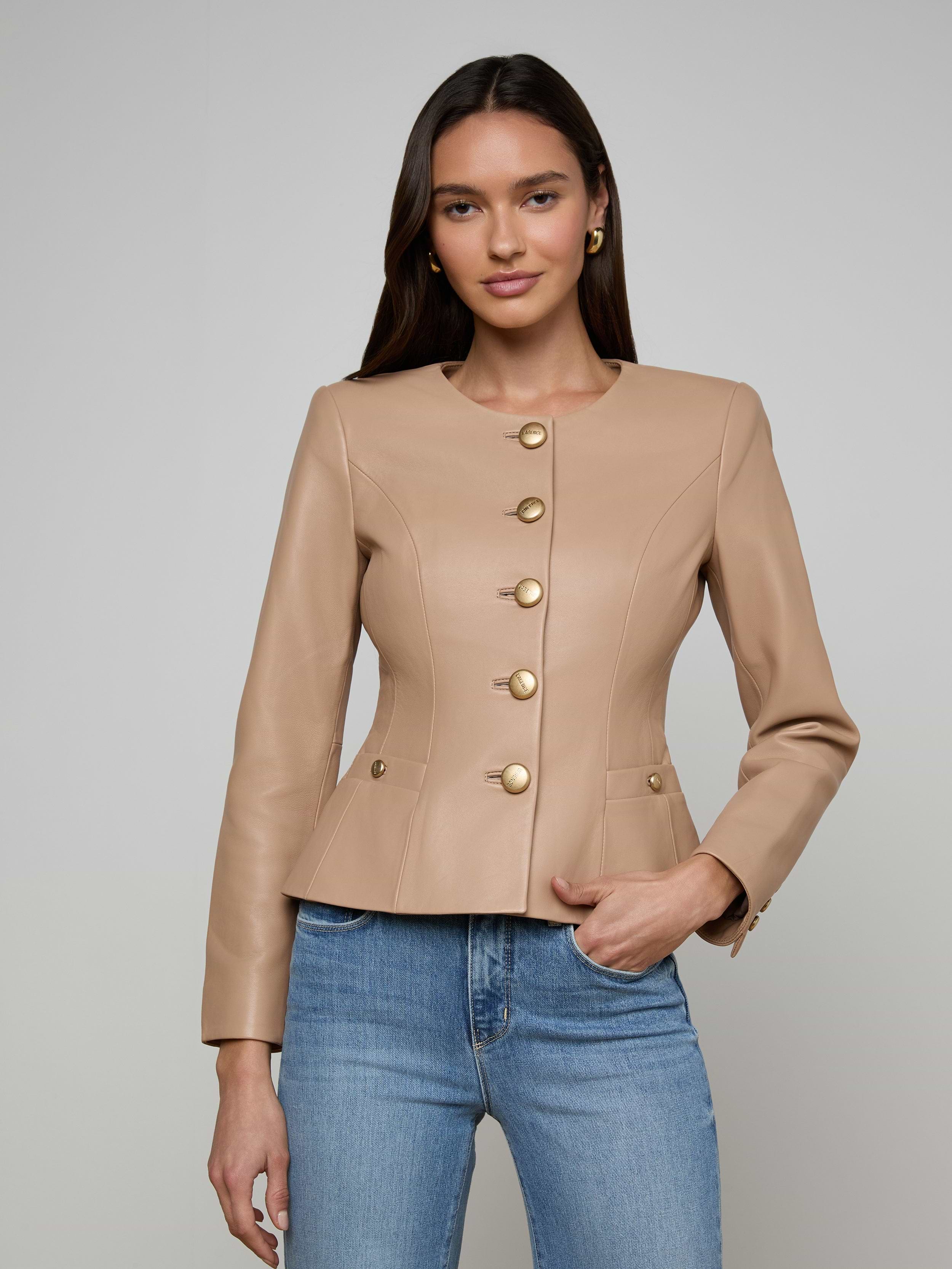 A woman wearing the Mariella Leather Jacket—a fitted, camel-colored chic leather piece with gold buttons—paired with blue jeans, stands against a plain, light background with a neutral expression.