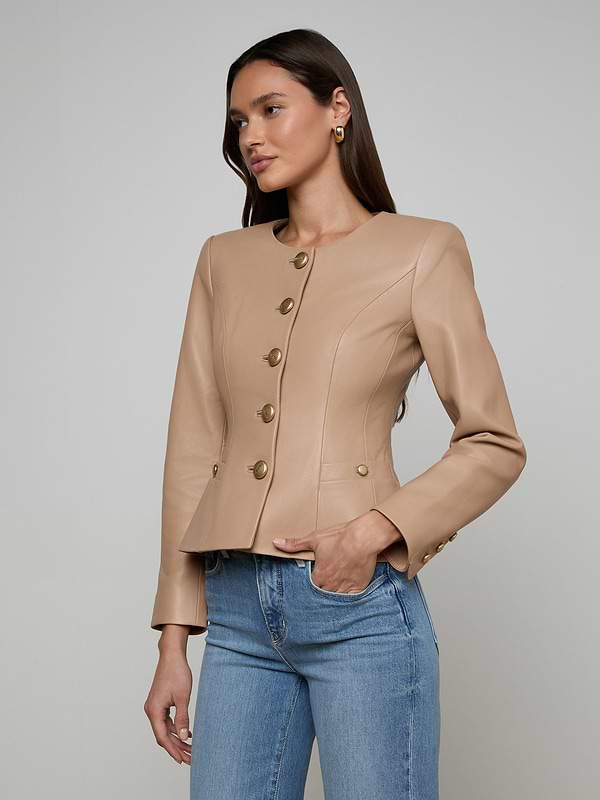 A woman models the Mariella Leather Jacket in a chic collarless design and cappuccino shade, styled with blue jeans. She stands against a plain light background, gazing slightly to the side with one hand in her pocket.