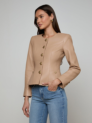 A woman models the Mariella Leather Jacket in cappuccino—fitted, collarless, with gold buttons—paired with light blue jeans, standing with one hand in her pocket against a plain light background.