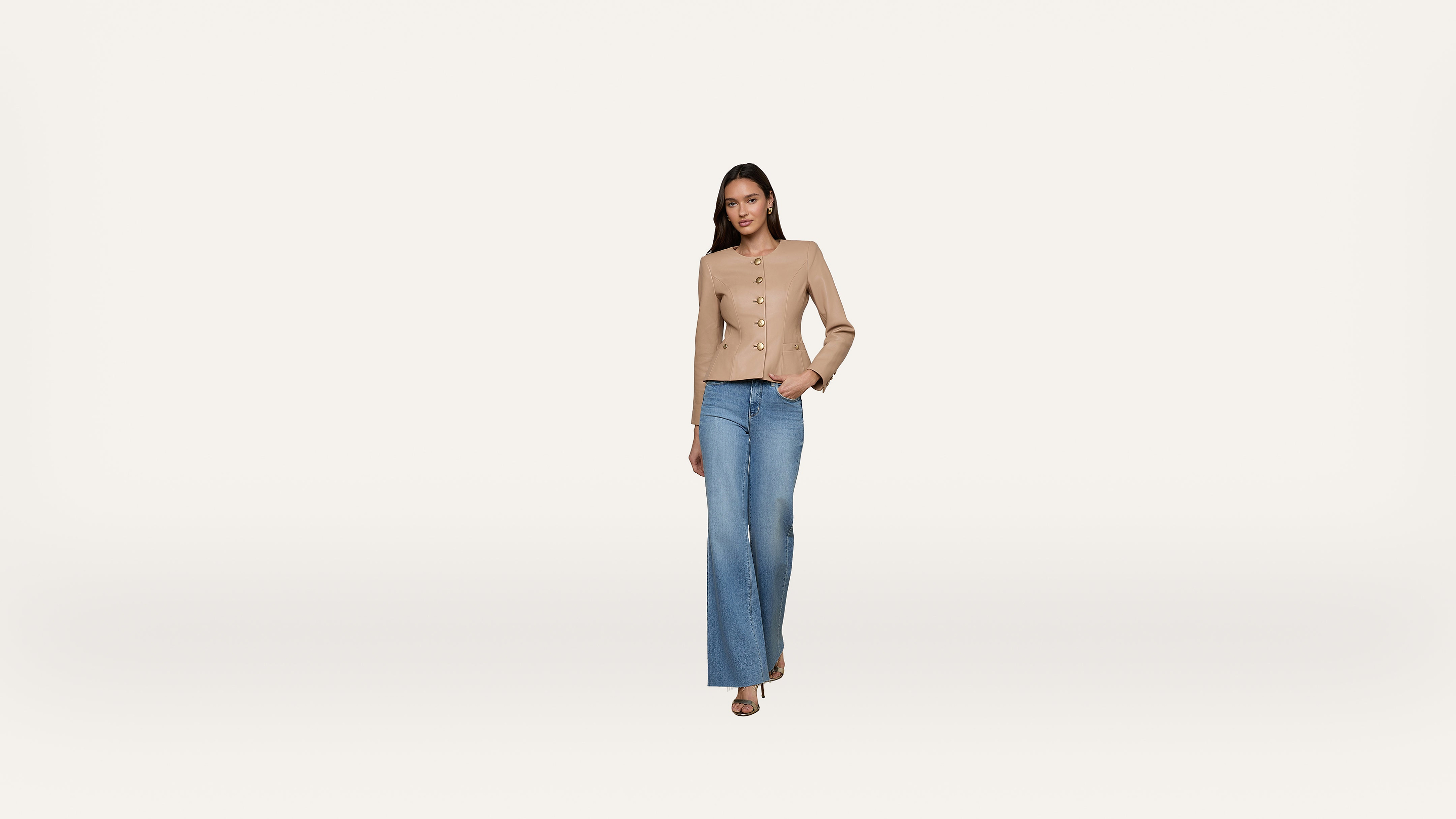 Woman wearing a tan button-up jacket and blue wide-leg jeans stands against a plain white background. She has one hand in her pocket and looks confidently at the camera.