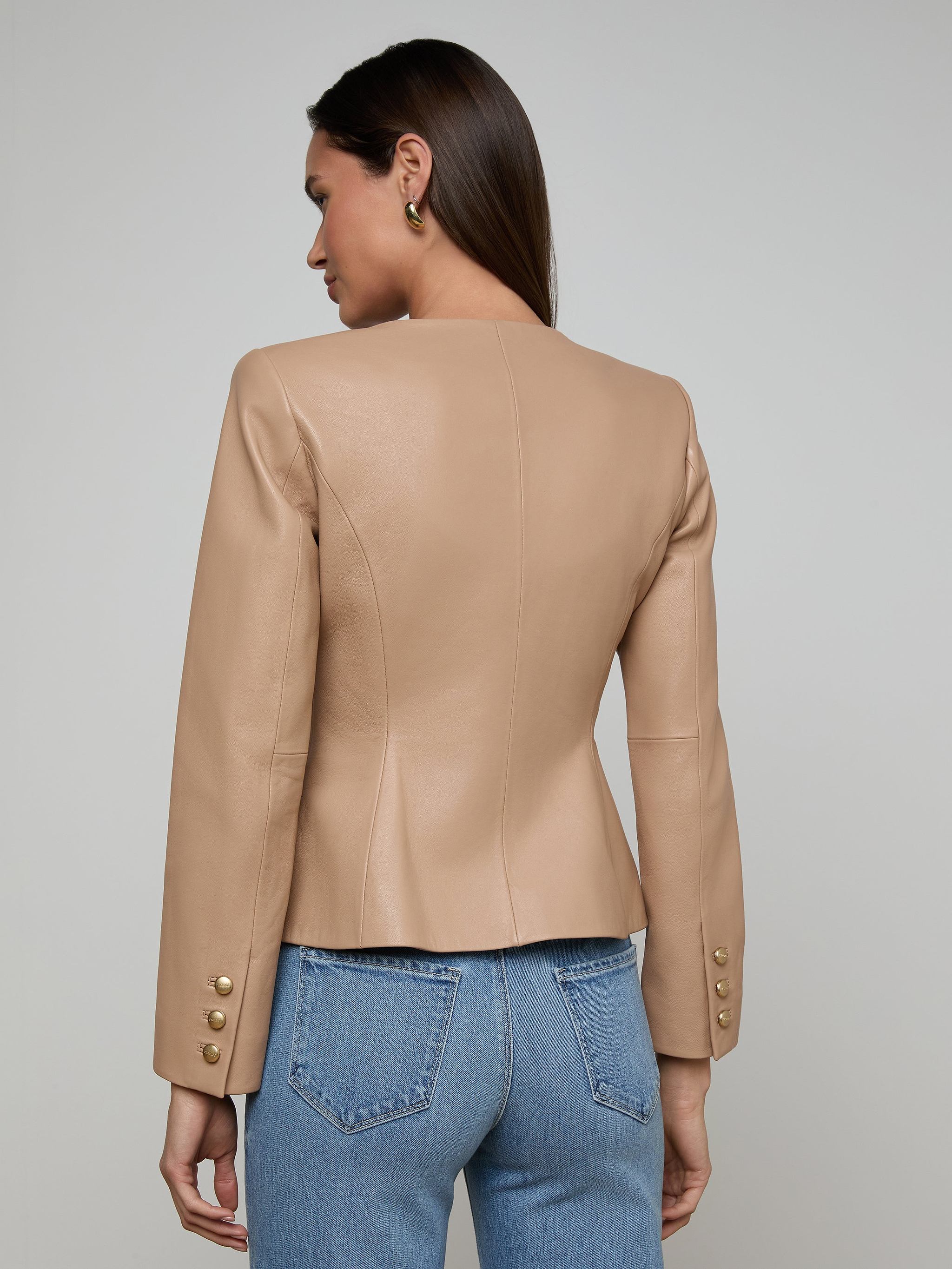 Wearing the Mariella Leather Jacket—designed without a collar and featuring button details on the sleeves—a woman in blue jeans stands with her back to the camera against a simple light background.