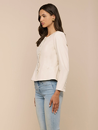 A woman with long brown hair stands sideways against a plain light background, styled in a cream-colored, button-up long-sleeve blouse and light blue jeans. She accessorizes with gold earrings and completes her outfit with the Mariella Leather Jacket for a chic finish.
