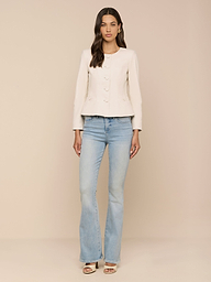 A woman stands against a plain white background, wearing the Mariella Leather Jacket over a light-colored long-sleeve top, paired with light blue flared jeans and open-toe heels. Her long wavy hair drapes over her shoulders.