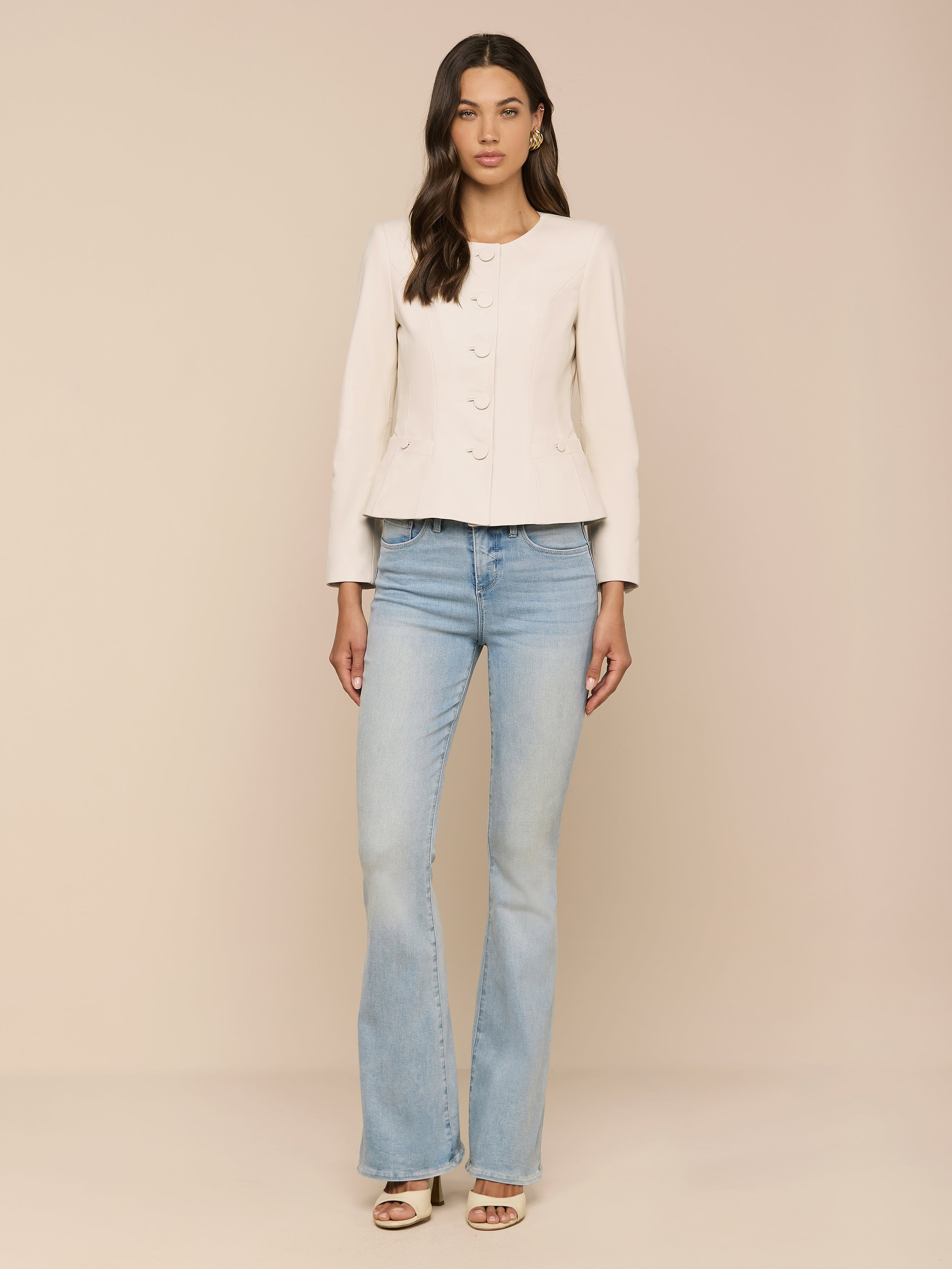 A woman stands against a plain white background, wearing the Mariella Leather Jacket over a light-colored long-sleeve top, paired with light blue flared jeans and open-toe heels. Her long wavy hair drapes over her shoulders.