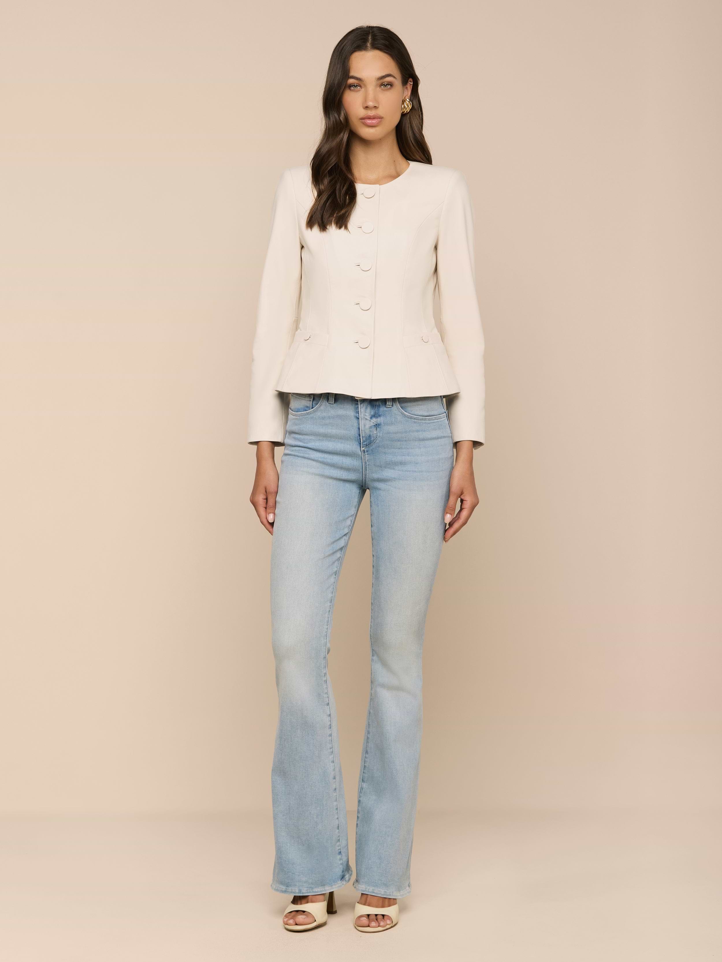 A woman stands against a plain white background, wearing the Mariella Leather Jacket over a light-colored long-sleeve top, paired with light blue flared jeans and open-toe heels. Her long wavy hair drapes over her shoulders.