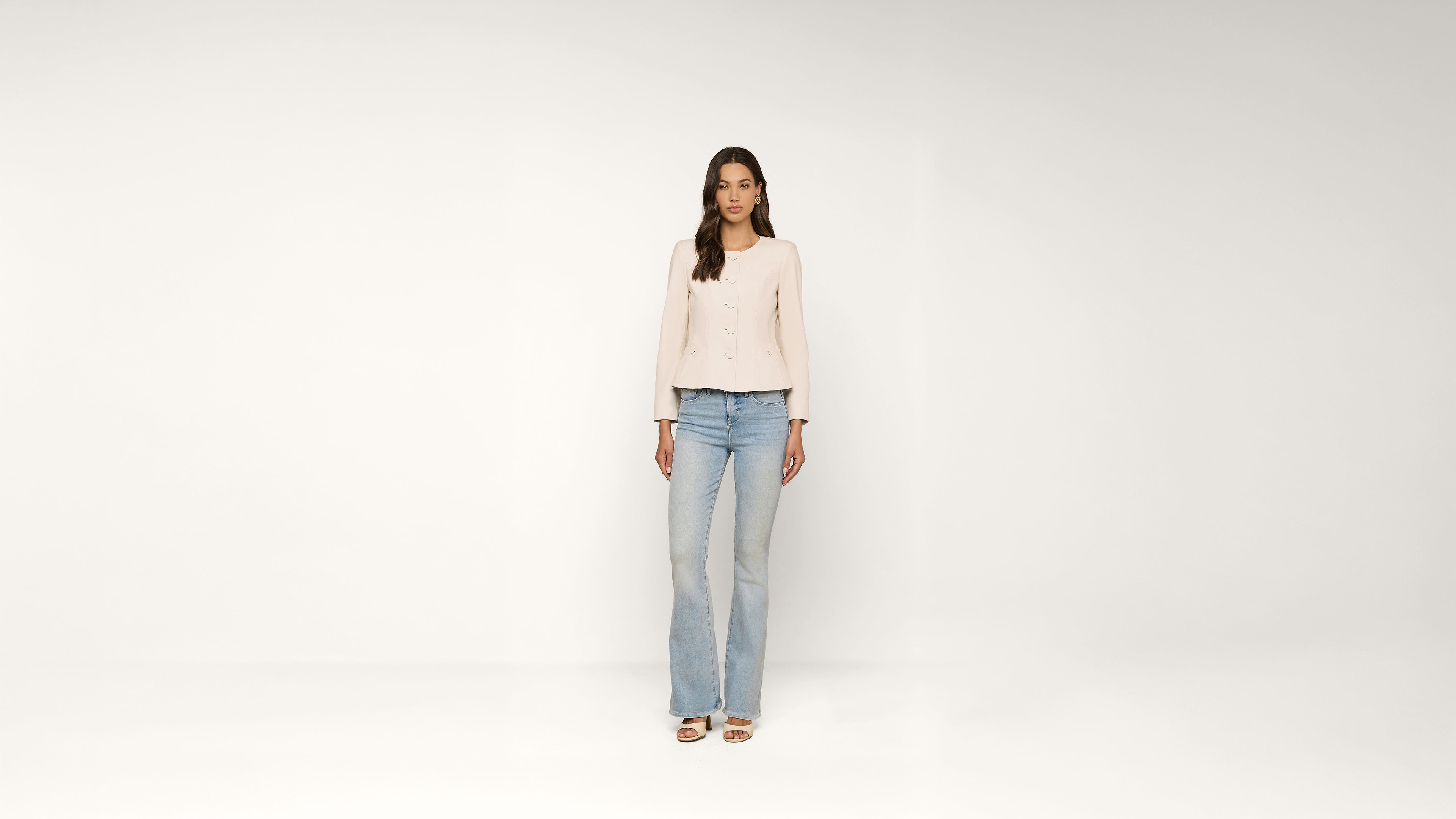Woman stands against a plain white background, wearing a light-colored blouse, blue jeans, and white open-toe shoes. She has long brown hair and her arms rest naturally by her sides.