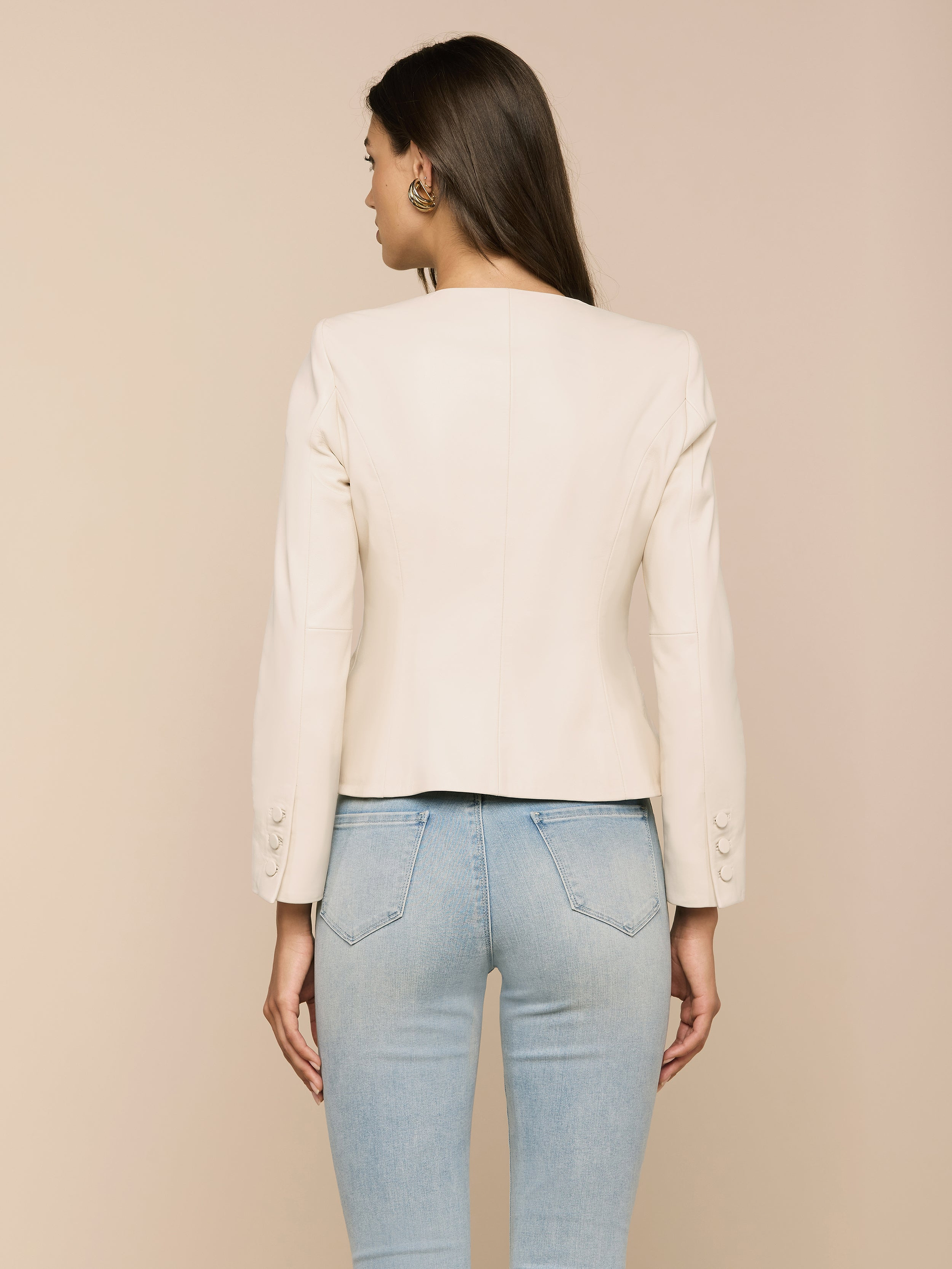 A woman facing away from the camera models the Mariella Leather Jacket—a chic, cream-colored, collarless design—paired with light blue jeans against a plain white background.