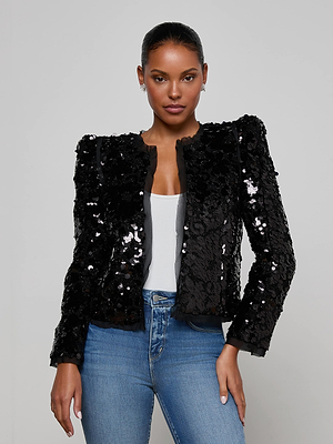 A woman models the Shai Paillette Jacket by L'AGENCE—a black cropped party jacket—styled over a white top and blue jeans, standing against a plain light gray background and gazing confidently at the camera.