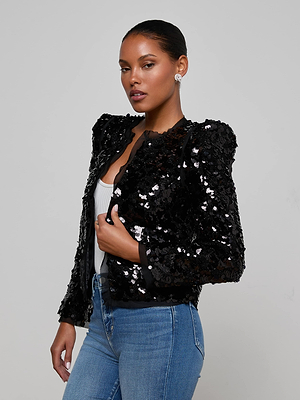 A woman with slicked-back hair models the L'AGENCE Shai Paillette Jacket over a white top and blue jeans, posing confidently against a plain gray background.