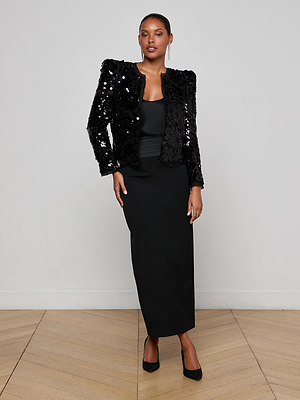 A woman stands on a wooden floor against a white background, wearing the L'AGENCE Shai Paillette Jacket over a black top, long black skirt, and heels. The chic cropped party jacket adds sparkle to her sophisticated look.