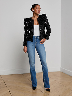 A woman poses indoors against a white wall and wooden floor, wearing the L'AGENCE Shai Paillette Jacket over a white top, paired with blue high-waisted jeans and black heels, one hand resting on her hip.