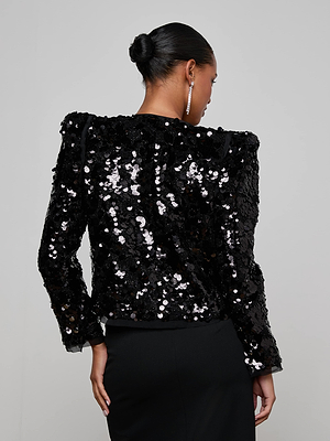 A woman with her hair in a neat bun and drop earrings stands with her back to the camera, wearing the L'AGENCE Shai Paillette Jacket paired with a black skirt for a chic, shimmering look.