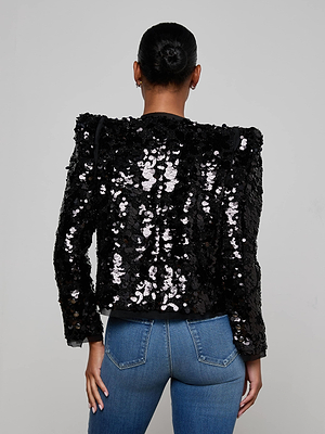 A person with dark hair in a bun, wearing the L'AGENCE Shai Paillette Jacket and blue jeans, stands facing away from the camera against a plain light background. The shimmering jacket adds a festive touch to the look.
